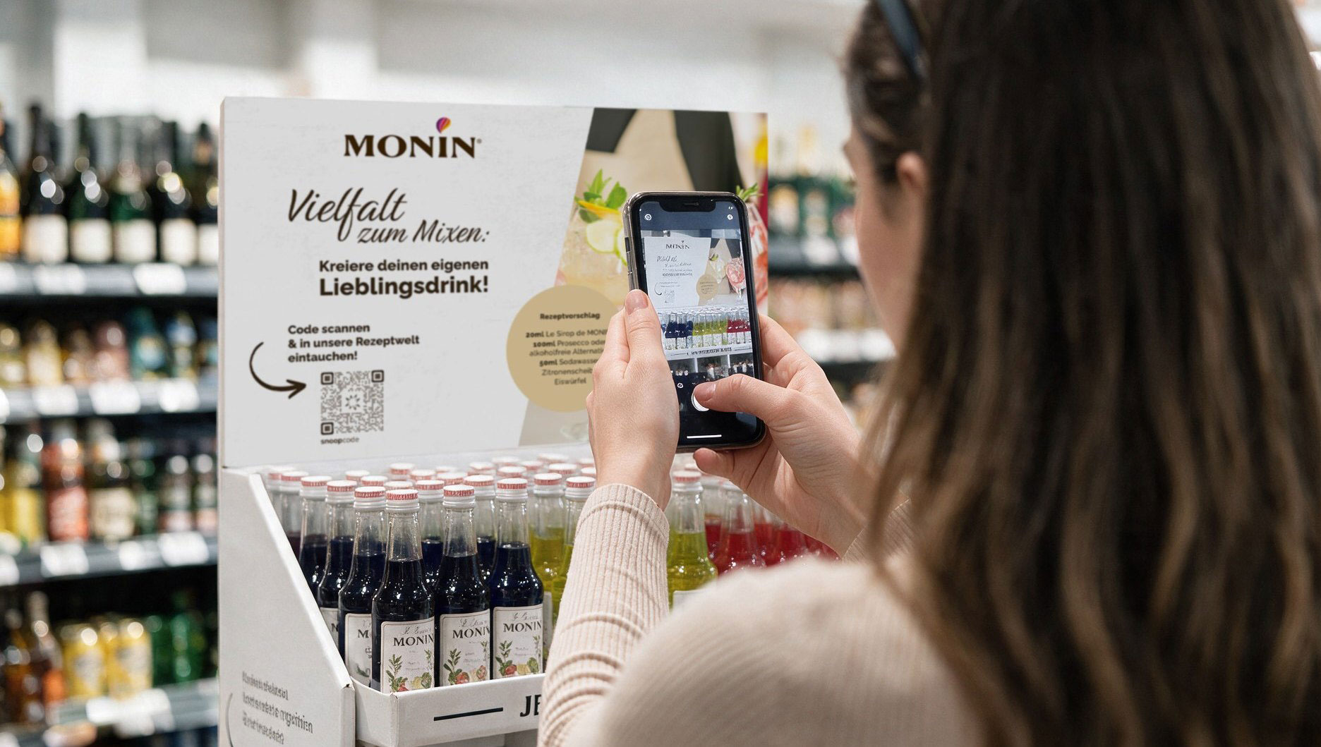 MONIN POS-Display im Handel: eine Kundin steht vor dem Aufsteller mit den Sirupflaschen und scannt mit dem Smartphone den 2D-Code auf dem Topschild. Auf dem Schild steht &bdquo;Vielfalt zum Mixen &ndash; kreiere deinen eigenen Lieblingsdrink&ldquo;.